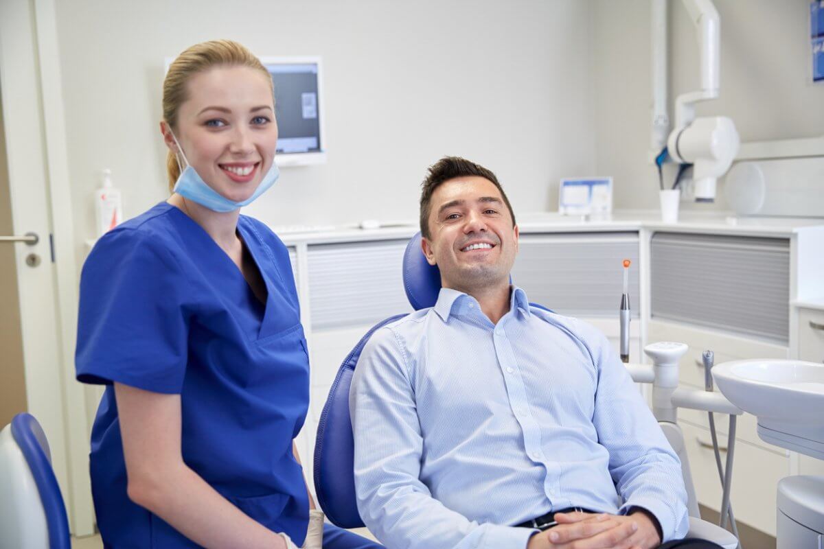 Professional man in a button-up shirt sitting in a dentists chair smiling next to a female dental hygienist dressed in blue