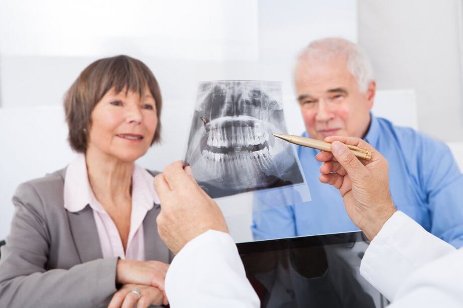 A dentist is holding up an x-ray and pointing to it, talking to an older male and female.