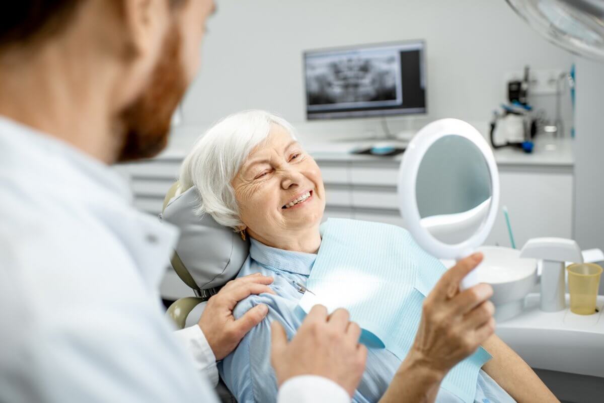 Older woman sitting in a dentists chair looking at herself in a handheld mirror & smiling as the dentist sits beside her.