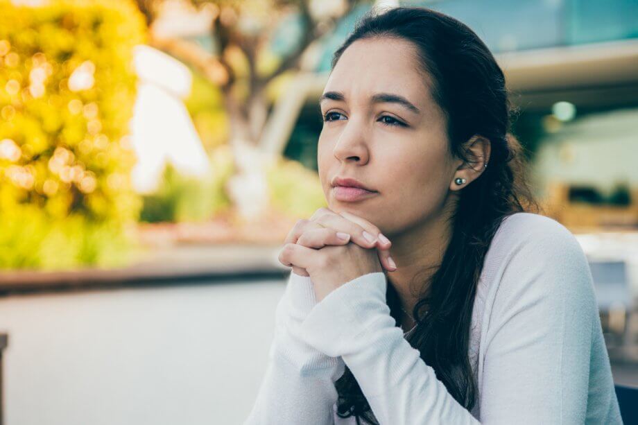 Woman looking contemplative w/ long dark hair sitting outdoors with her hands under her face looking off into the distance