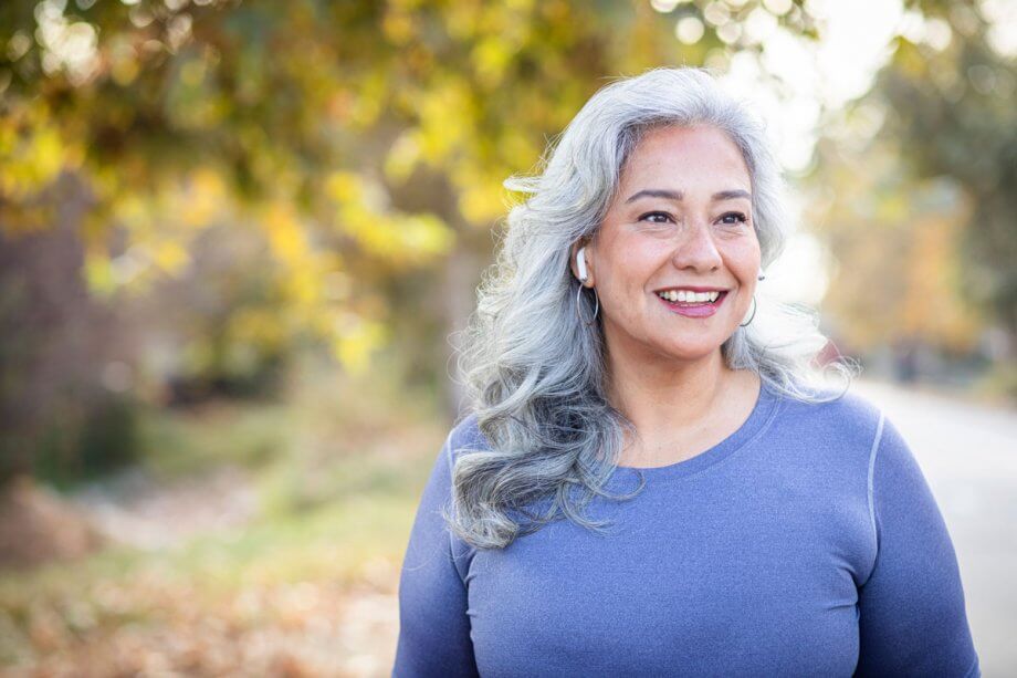 older woman with ear buds smiling, standing outside