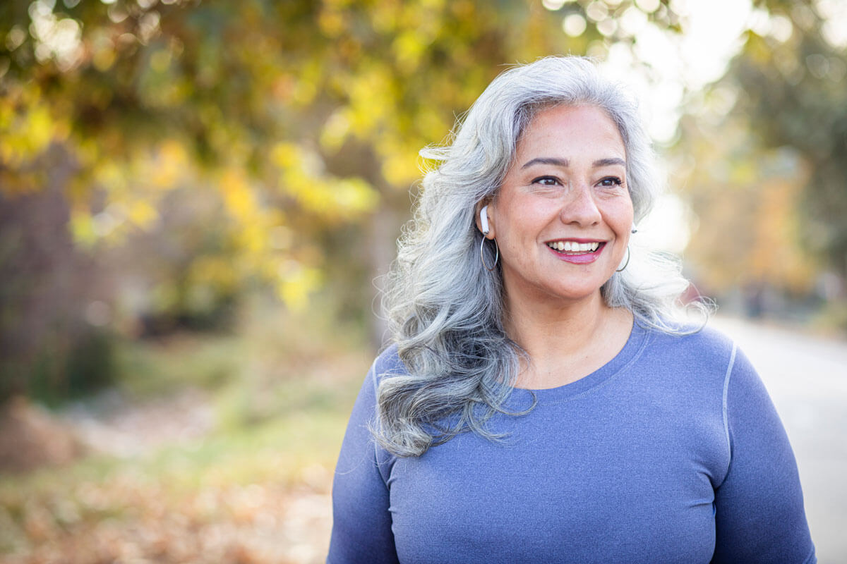 older woman with ear buds smiling, standing outside